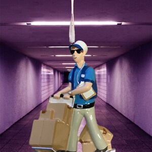 May include: A blue and white Christmas ornament depicting a delivery person pushing a cart with boxes. The ornament is in a hallway with purple walls. The ornament has the text "Pete 2.02.3" on the base.