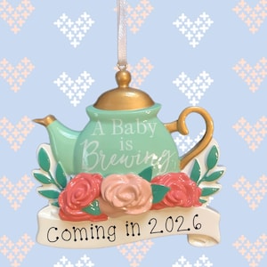 May include: A decorative ornament shaped like a teapot, with a gold lid and handle, and a mint green body. The text "A Baby is Brewing" is on the teapot. Below are pink and peach roses and the text "Coming in 2026" on a banner.