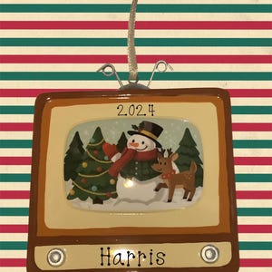 May include: A brown and beige retro television ornament with a snowman and reindeer scene inside the screen. The ornament has a white knob on each side and the year 2024 printed on the top. The name "Harris" is printed on the bottom of the ornament.
