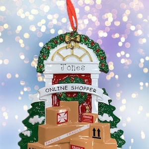 May include: A Christmas ornament shaped like a house with a wreath on the door. The house has a red door with the name "Jones" on it and the words "ONLINE SHOPPER" on a banner above the door. The house is surrounded by boxes labeled "FRAGILE" and "HANDLE WITH CARE".