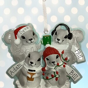 以下が含まれることがあります： 冬帽とマフラーをつけた4匹の灰色のリスが描かれたクリスマスオーナメント。リスたちはコーヒーカップ、イルミネーション、ヘッドホンを持っています。オーナメントには「Happy Holidays」の文字と「Dave」「Adam」「Bill」「Phil」の名前が書かれています。