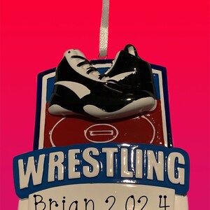 May include: A white and blue wrestling ornament with a red wrestling mat and black wrestling shoes. The ornament has the text "WRESTLING" and "Brian 2024".