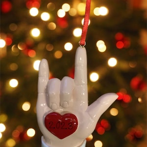 May include: White ceramic Christmas ornament shaped like a hand signing 'I love you' in American Sign Language. The ornament has a red heart with the year 2023 and the name Katrina written on the bottom.