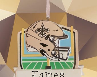 May include: A football-themed ornament featuring a tan helmet with a fleur-de-lis emblem, set against a backdrop of a goalpost and field. The name "James" and the year "2.025" are displayed on a banner below. A light pink ribbon is attached for hanging.