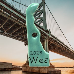 May include: A light blue ceramic ornament shaped like the state of Delaware with the year 2023 and the word "Wes" written on it. The ornament is in front of a large bridge.