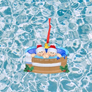 Könnte beinhalten: Ein Weihnachtsbaumschmuck mit zwei Personen in einem Whirlpool mit einer Gummiente. Der Schmuck ist aus Keramik und in realistischer Art bemalt. Der Whirlpool ist braun und hat einen weißen Rand. Die Personen tragen Weihnachtsmützen und haben lächelnde Gesichter. Der Schmuck hängt an einem roten Band.