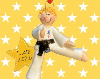 May include: A ceramic ornament of a child in a karate uniform, with a black belt and a star-shaped name tag that reads "Liam 2025." The ornament has a red ribbon for hanging and is set against a yellow background with white stars.