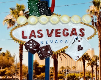 May include: A white and gold Las Vegas sign with red glitter lettering. The sign has a stack of chips, dice, and a playing card with an ace of hearts on it. The sign says "Las Vegas, Nevada".