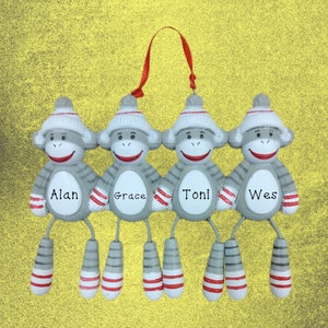 May include: Four gray sock monkey ornaments with red and white stripes. The monkeys are hanging together and each has a name written on their chest: Alan, Grace, Toni, and Wes.