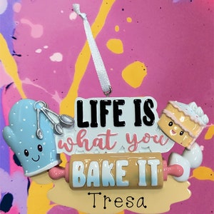 Könnte beinhalten: Ein weißer und blauer Weihnachtsbaumschmuck mit einem Nudelholz, einem blauen Ofenhandschuh und einem Kuchen. Der Schmuck sagt "Life is what you bake it" und "Tresa".