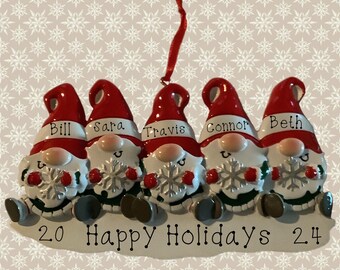 May include: Five white gnomes with red hats and white beards are holding snowflakes. The gnomes are sitting in a row and have the names Bill, Sara, Travis, Connor, and Beth written on their hats. The gnomes are on a white background with the text "2.0 Happy Holidays 2.4".