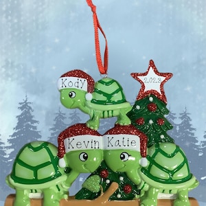 May include: A Christmas ornament featuring a family of green turtles wearing Santa hats. The ornament includes the names "Kody", "Kevin", and "Katie", along with a star with the year "2023" and the text "Merry Christmas".