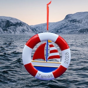 May include: Red, white and blue nautical themed Christmas bauble with a sailboat in the centre. The bauble is shaped like a life preserver and has the year 2023 and the name James written on it.