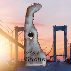 May include: A ceramic ornament shaped like the state of Delaware with the year 2023 and the name Shane written on it. The ornament is in front of a bridge with a sunset in the background.