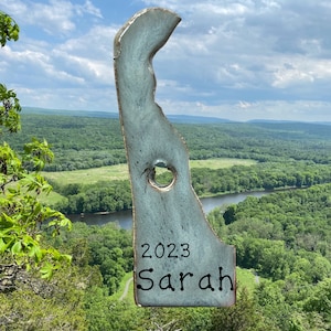 May include: A light blue ceramic state-shaped ornament with a hole in the middle. The ornament is decorated with the year 2023 and the name Sarah.