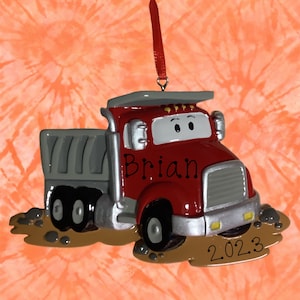 Könnte beinhalten: Roter und grauer Kipper-Weihnachtsbaumschmuck mit dem Namen "Brian" und dem Jahr "2023". Der LKW befindet sich in einer Pfütze.