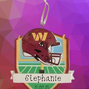 May include: A burgundy football helmet ornament with a gold "W" logo, set against a shield design with a green field and blue sky. The name "Stephanie" and the year "2025" are displayed on a white banner.