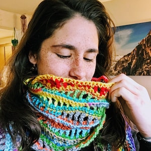 May include: A woman wearing a colorful crocheted scarf with a multi-colored striped pattern. The scarf is made of yarn in shades of red, orange, yellow, green, blue, and purple.