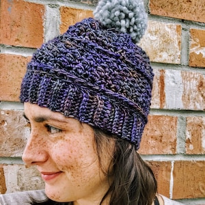 May include: A purple and blue crocheted beanie with a gray pom pom on top. The beanie is worn by a person with freckles.