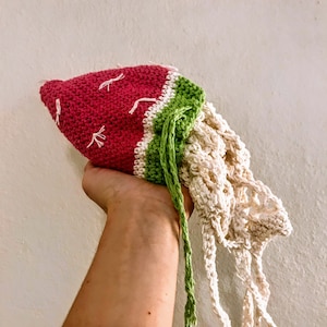 May include: A handmade crochet bag in the shape of a watermelon. The bag features a red top with white seed details, a green rind, and a cream-colored net-like bottom. A green drawstring is attached.