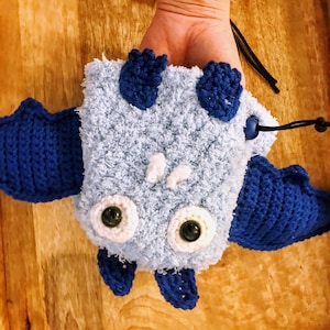 May include: A handmade, light blue and dark blue crocheted bat-shaped pouch with large, round eyes and small white teeth. The pouch has dark blue wings and feet, and a black drawstring closure.