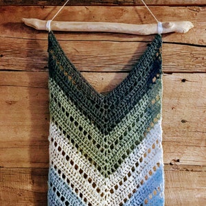 Peut inclure: Une tapisserie murale en crochet avec un motif en chevron dans des tons de bleu, vert et blanc. La tapisserie est fixée à une branche de bois naturel.