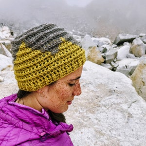 Puede incluir: Una mujer con un gorro de punto amarillo y gris. El gorro tiene un dise&ntilde;o texturizado y se lleva sobre la cabeza. Est&aacute; de pie frente a un fondo rocoso.