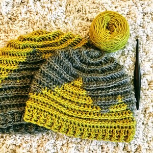 Puede incluir: Un gorro de crochet con un patr&oacute;n de color gris y amarillo. El gorro est&aacute; hecho con un punto texturizado y tiene un ala ancha. El gorro est&aacute; sobre una superficie blanca y esponjosa.
