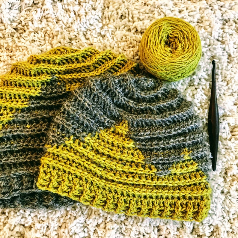 Puede incluir: Un gorro de crochet con un patr&oacute;n de color gris y amarillo. El gorro est&aacute; hecho con un punto texturizado y tiene un ala ancha. El gorro est&aacute; sobre una superficie blanca y esponjosa.