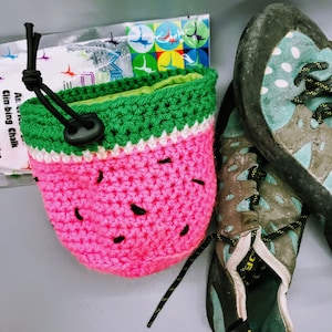 May include: A crocheted watermelon-shaped pouch with a green and white trim. The pouch is sitting on a gray surface next to a pair of brown and blue sneakers.