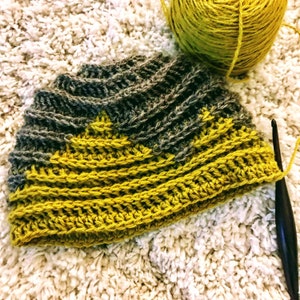Puede incluir: Un gorro de crochet con un patr&oacute;n de rayas amarillas y grises. El gorro se est&aacute; trabajando con un gancho de crochet.