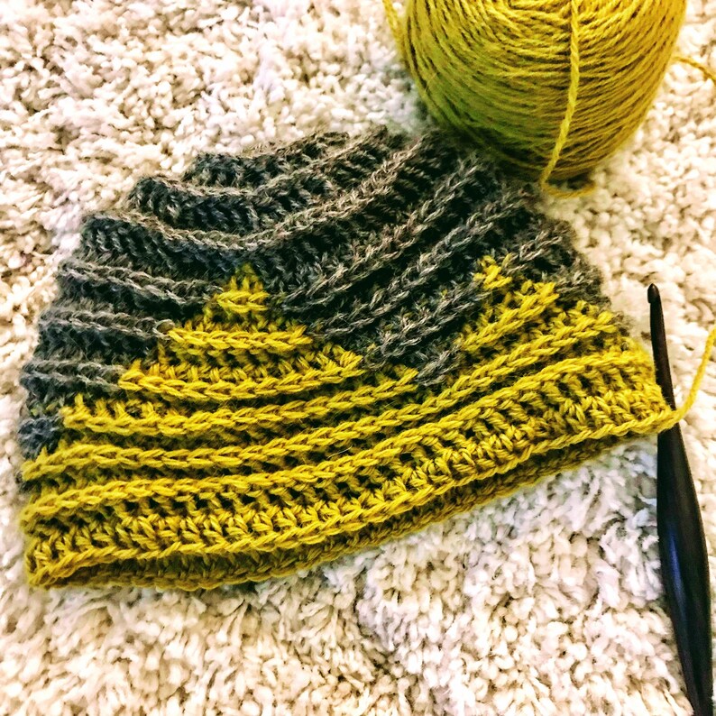 Puede incluir: Un gorro de crochet con un patr&oacute;n de rayas amarillas y grises. El gorro se est&aacute; trabajando con un gancho de crochet.
