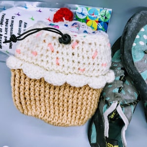 May include: A crocheted cupcake-shaped pouch with a white and brown design. The pouch has a pink and white frosting design and a red cherry on top. The pouch is sitting next to a pair of blue and black climbing shoes.