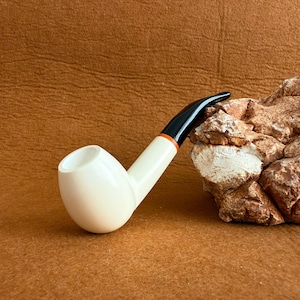May include: A white tobacco pipe with a black stem and orange accent. The pipe is set against a brown background and a textured rock formation. This pipe is designed for smoking tobacco.