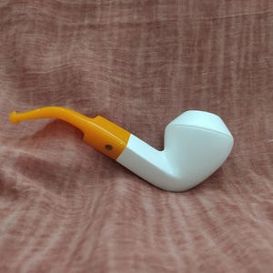 May include: A white pipe with an orange stem and accents. The pipe has a classic shape with a rounded bowl and a curved stem. The orange and white color combination gives the pipe a modern look. This smoking pipe is a unique gift.