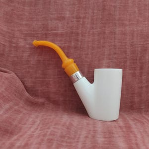 May include: A white ceramic pipe with an orange mouthpiece. The pipe is sitting on a pink fabric surface.