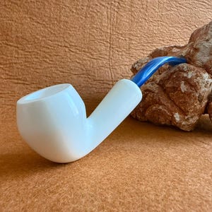 May include: A white pipe with a blue stem rests against a textured, brown rock formation. The pipe has a curved design with a rounded bowl and a cylindrical stem. The background is a solid, light brown color.