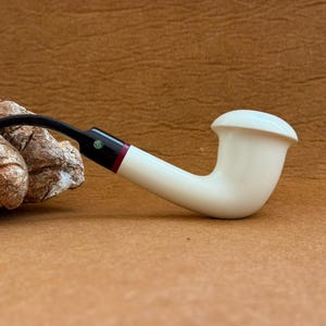 May include: A cream-colored tobacco pipe with a curved stem and a rounded bowl. The stem is black with a red and white accent near the mouthpiece. The pipe rests on a brown surface.