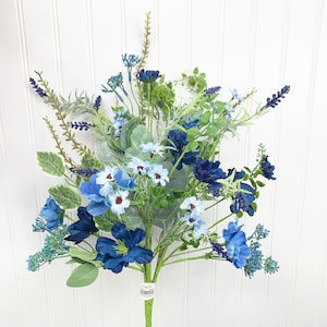 May include: A bouquet of artificial flowers in shades of blue and green. The arrangement includes various flower types, such as hydrangeas and daisies, with green foliage. The stems are wrapped in green tape. The background is a white, vertical-slat wall.