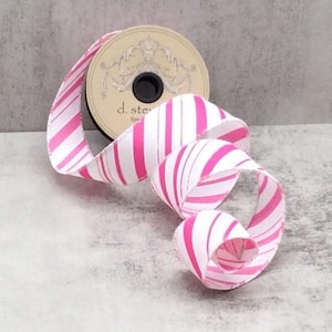 May include: A roll of white ribbon with pink stripes, partially unrolled, creating a looping pattern. The ribbon is on a neutral background. The spool has a beige label with decorative elements and the text "d. ste".
