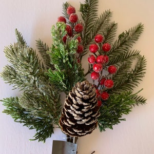 May include: A frosted pine sprig with red berries and a brown pine cone. The sprig is tied with twine and has a gray tag.