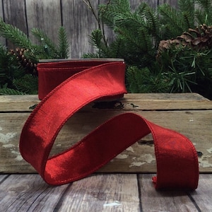 May include: A roll of red ribbon with a textured, shimmering surface. The ribbon is displayed on a wooden surface with evergreen branches and pine cones in the background. The ribbon is approximately 2 inches wide.