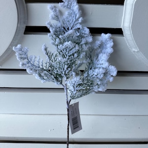 May include: A decorative artificial evergreen branch featuring dark green foliage heavily coated in a thick layer of white, snow-like texture. The stem is brown and includes a small white rectangular product tag with a barcode attached near the base.