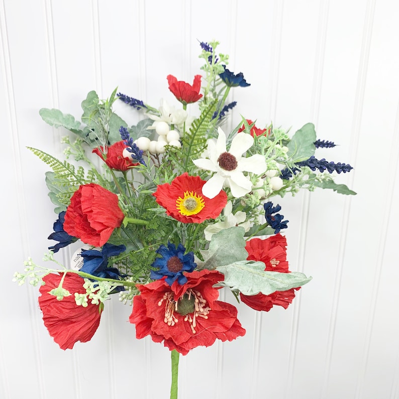 May include: A vibrant floral arrangement featuring red poppies, white daisies, and blue accents. The bouquet includes green foliage and white berries, creating a patriotic color scheme. The flowers are arranged on a green stem.