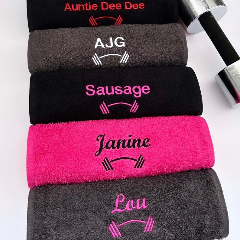 May include: Five personalized towels with names embroidered in different colors. The towels are stacked on top of each other. Each towel has a dumbbell graphic embroidered on it. The names on the towels are Auntie Dee Dee, AJG, Sausage, Janine, and Lou.