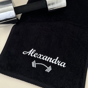 May include: Black gym towel with white embroidery. The embroidery says "Alexandra" with a barbell symbol.