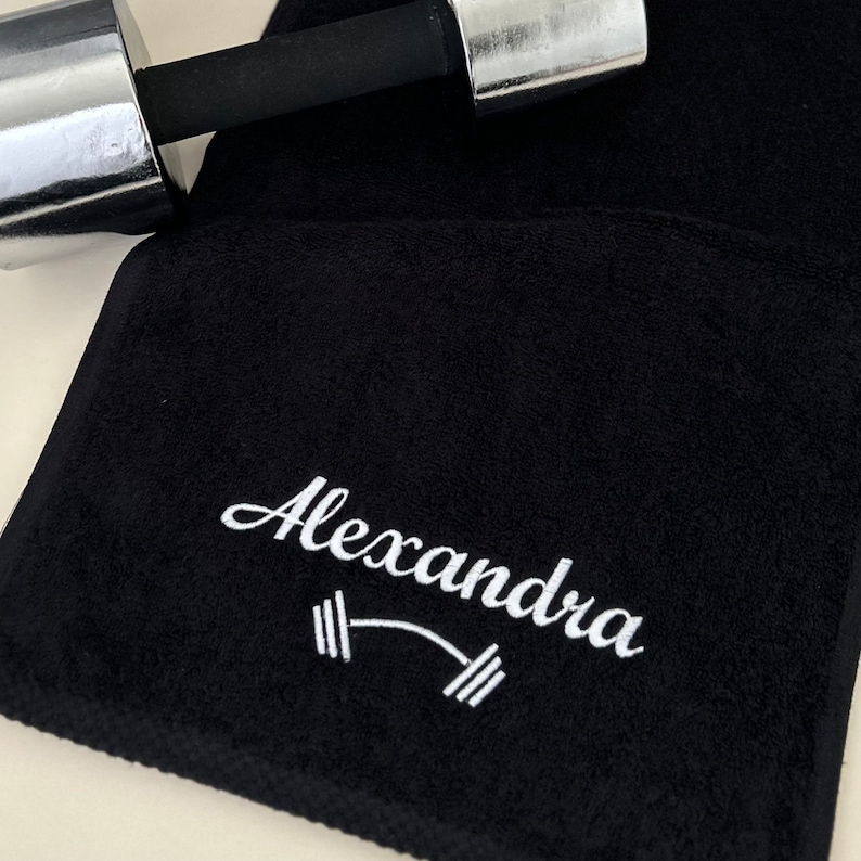 May include: Black gym towel with white embroidery. The embroidery says "Alexandra" with a barbell symbol.