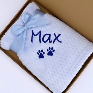 Op de afbeelding: Een lichtblauwe babydeken met een blauw lint en de naam "Max" erop geborduurd. Twee blauwe pootjes zijn ook geborduurd op de deken.