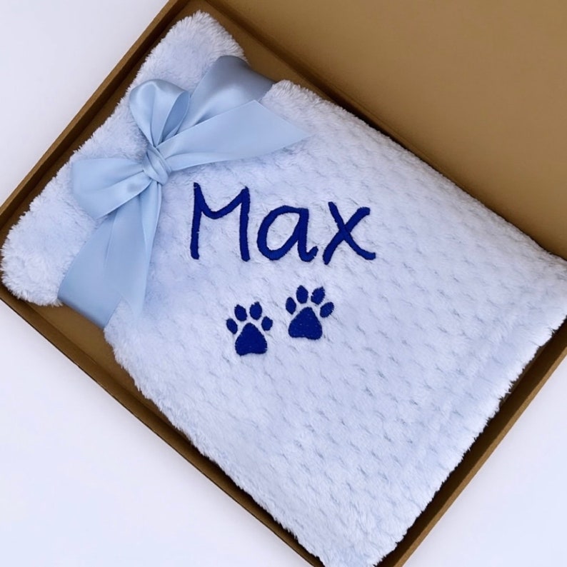 Op de afbeelding: Een lichtblauwe babydeken met een blauw lint en de naam "Max" erop geborduurd. Twee blauwe pootjes zijn ook geborduurd op de deken.