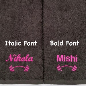 May include: Two dark gray towels with pink embroidery. The left towel has the name "Nikola" in italic font and a pink dumbbell symbol. The right towel has the name "Mishi" in bold font and a pink dumbbell symbol.